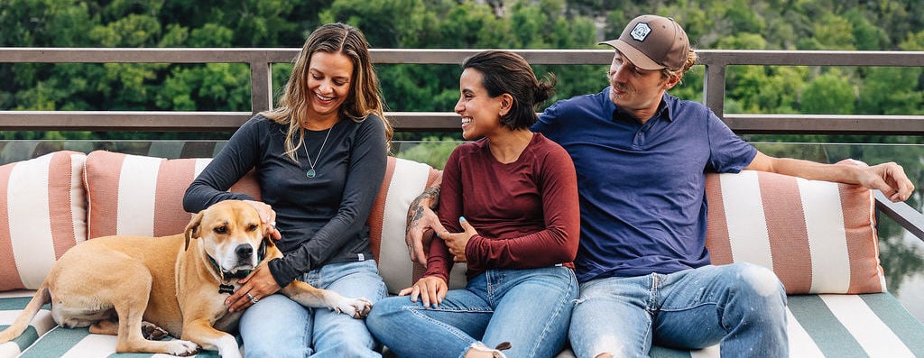 A lifestyle photo from Whole Earth's private label photoshoot. It features a man and two women sitting on an outdoor couch on a patio and they're all wearing Whole Earth apparel. A large, yellow, short-haired dog is also sitting with them.