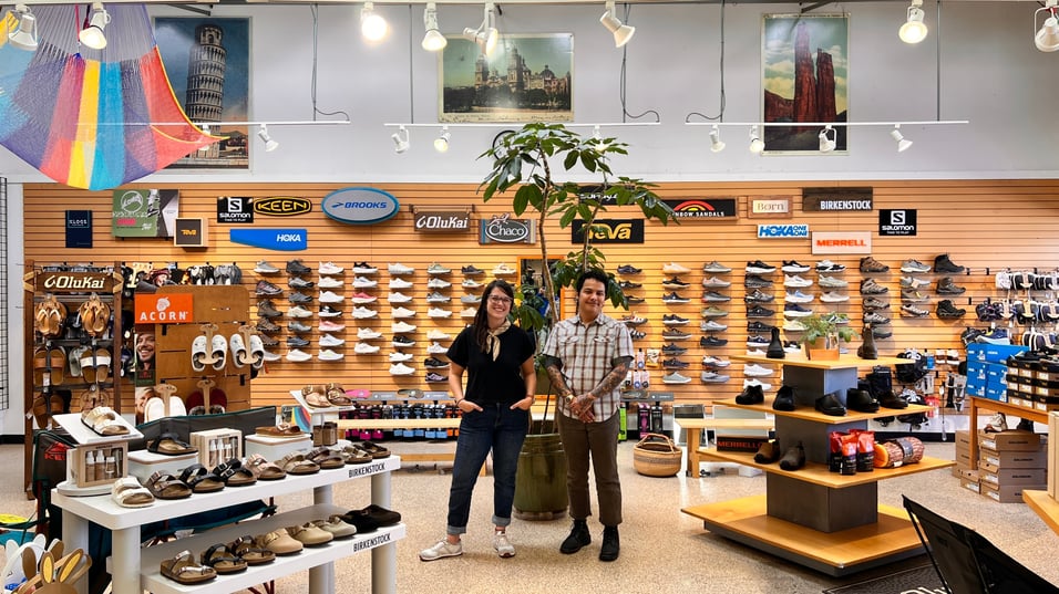 The new footwear buyers stand proudly in the shoe department. Lauren, a woman with dark brown mid length hair, stands smiling with her hands in her pockets to the left of TJ. He has short dark hair, and many tattoos.